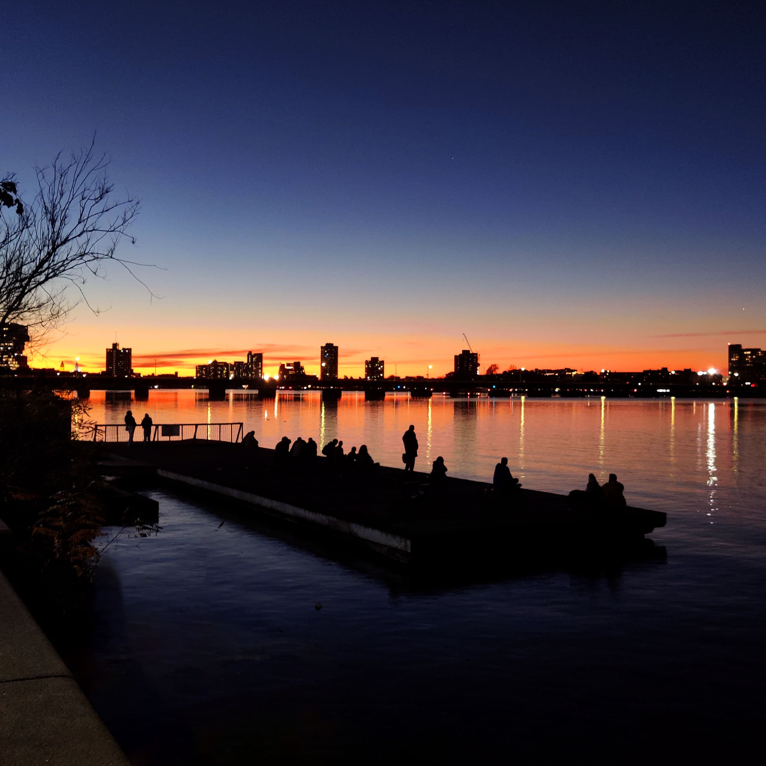 looking over the charles river at people watching the sunset on a public dock